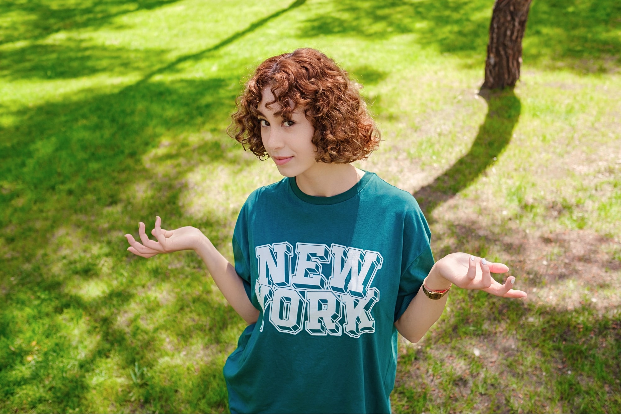 A person with curly hair is standing on a grassy area, wearing a green shirt with "New York" printed on it. They have an expression of uncertainty, with their hands raised and shoulders slightly shrugged. Shadows of trees are visible on the grass.