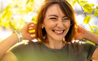 A woman with shoulder-length brown hair smiling and looking joyful, stands outdoors surrounded by bright green leaves. She touches her neck, wearing a green shirt and a white watch. Sunlight filters through the foliage, creating a warm atmosphere.
