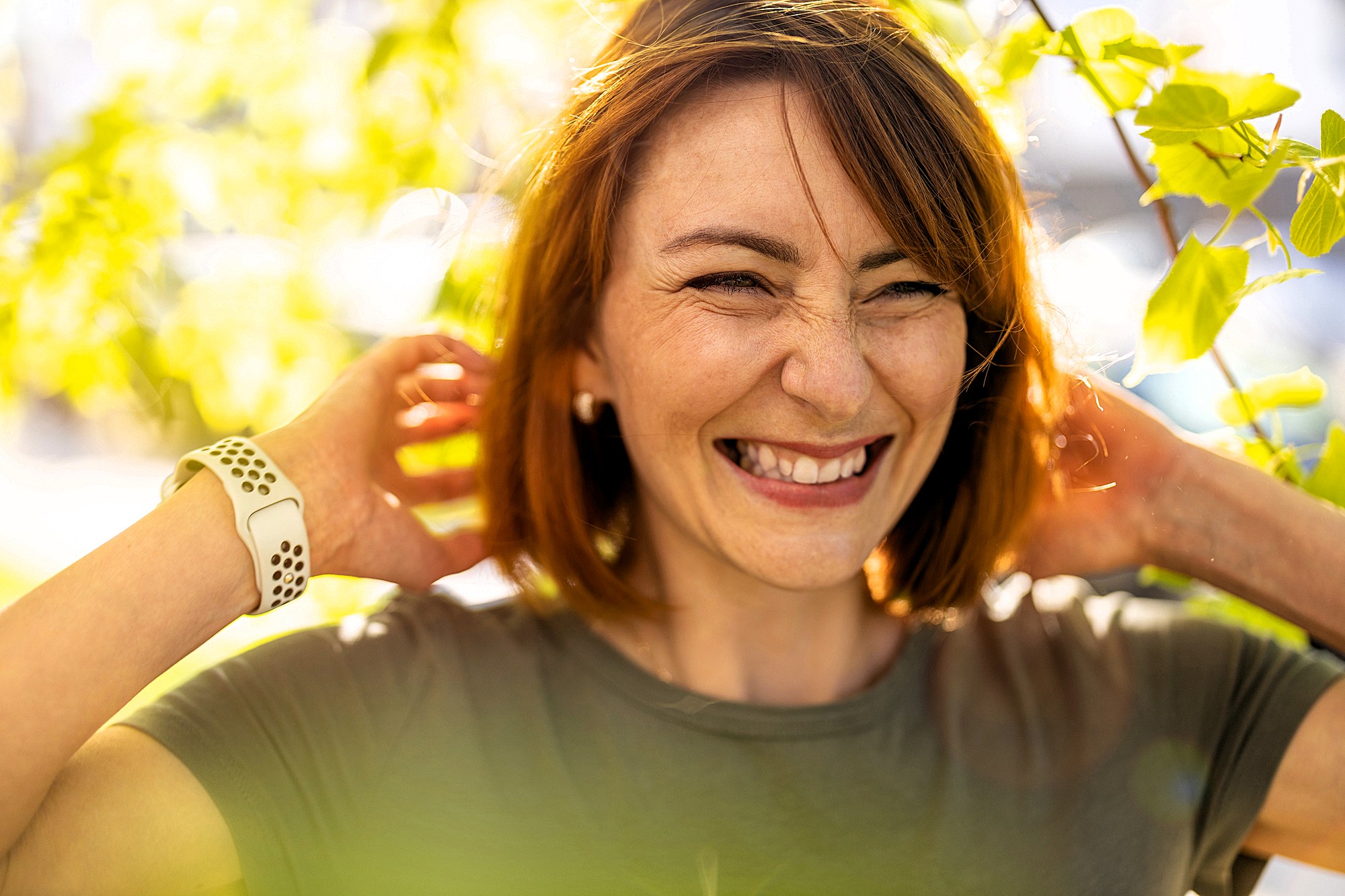 A woman with shoulder-length brown hair smiling and looking joyful, stands outdoors surrounded by bright green leaves. She touches her neck, wearing a green shirt and a white watch. Sunlight filters through the foliage, creating a warm atmosphere.