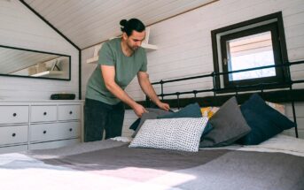 A person in a green shirt arranges pillows on a neatly made bed in a cozy, white-paneled attic bedroom with a small window and a dresser with a mirror.