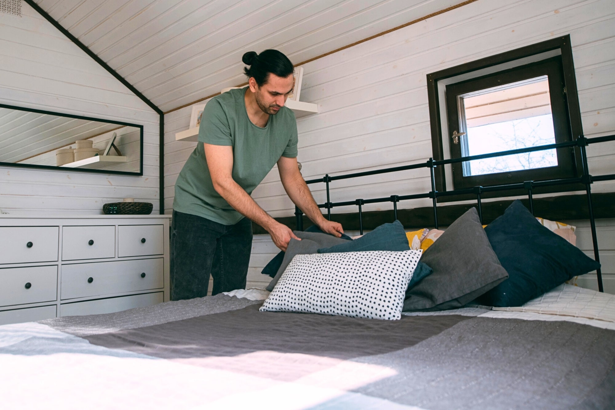 A person in a green shirt arranges pillows on a neatly made bed in a cozy, white-paneled attic bedroom with a small window and a dresser with a mirror.