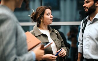 Three people are standing and talking near a glass wall. The woman in the center is holding a pen, while another person in the foreground holds a folder and phone. A man with a beard stands on the right, smiling.