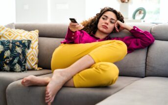 A woman in a pink shirt and yellow pants lounges on a sofa, holding a TV remote and looking bored or pensive. Patterned cushions are beside her, and she is barefoot with legs curled up.
