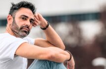 A man with short dark hair and a beard sits outdoors, wearing a white t-shirt and light blue jeans. He rests his arm on his knee and gazes thoughtfully at the camera. The background is blurred.