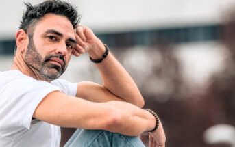 A man with short dark hair and a beard sits outdoors, wearing a white t-shirt and light blue jeans. He rests his arm on his knee and gazes thoughtfully at the camera. The background is blurred.