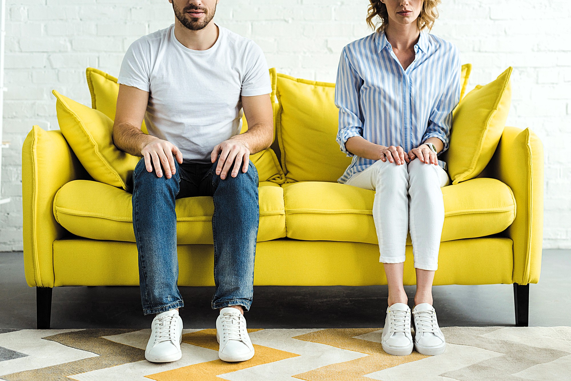 A man and woman sit on a bright yellow couch, both wearing white sneakers. The man wears a white T-shirt and jeans, while the woman wears a blue-striped shirt and white pants. They look straight ahead, not touching, against a white brick wall.