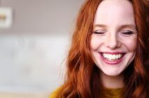 A woman with long red hair smiles with her eyes closed, showing her teeth. She is wearing a mustard yellow sweater and appears joyful. The background is softly blurred.