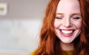 A woman with long red hair smiles with her eyes closed, showing her teeth. She is wearing a mustard yellow sweater and appears joyful. The background is softly blurred.