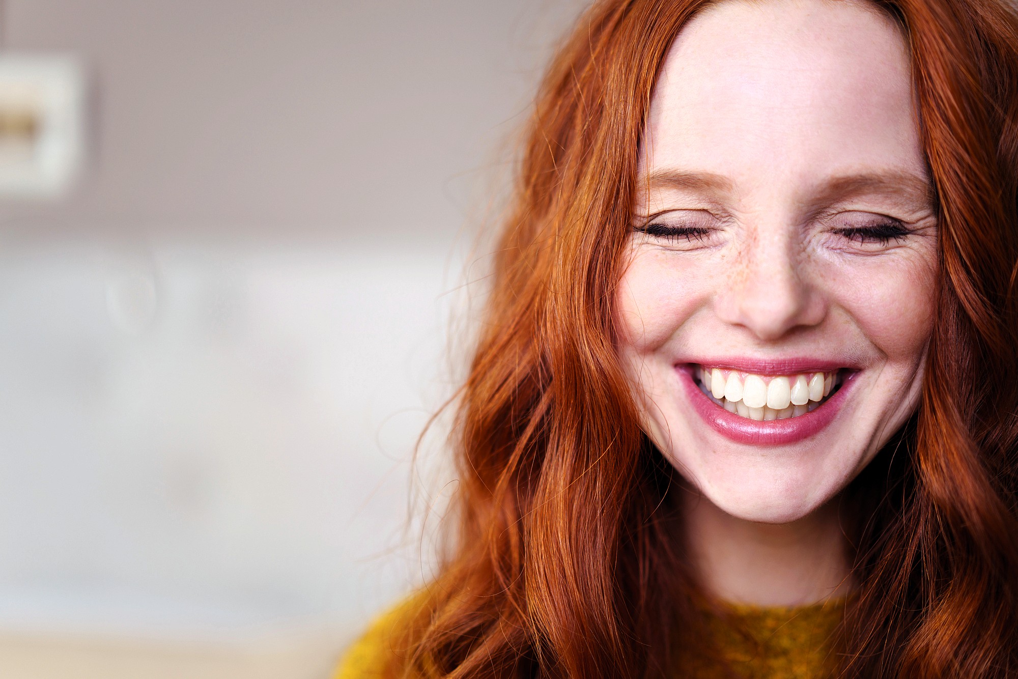A woman with long red hair smiles with her eyes closed, showing her teeth. She is wearing a mustard yellow sweater and appears joyful. The background is softly blurred.