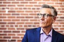 A mature man wearing glasses and a navy blazer over a lavender shirt stands against a brick wall, looking thoughtfully into the distance.