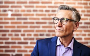 A mature man wearing glasses and a navy blazer over a lavender shirt stands against a brick wall, looking thoughtfully into the distance.