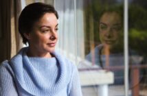 A woman wearing a light blue sweater gazes out a window, her reflection visible in the glass. She appears thoughtful, with short dark hair, as natural light illuminates her face.