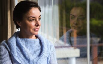 A woman wearing a light blue sweater gazes out a window, her reflection visible in the glass. She appears thoughtful, with short dark hair, as natural light illuminates her face.