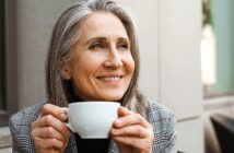 A smiling woman with long gray hair holds a white coffee cup, looking thoughtfully into the distance. She is wearing a plaid jacket and sitting outdoors, with a blurred building wall in the background.