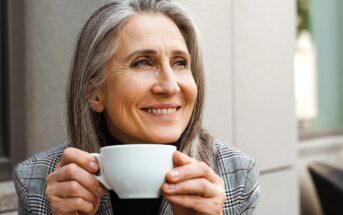 A smiling woman with long gray hair holds a white coffee cup, looking thoughtfully into the distance. She is wearing a plaid jacket and sitting outdoors, with a blurred building wall in the background.