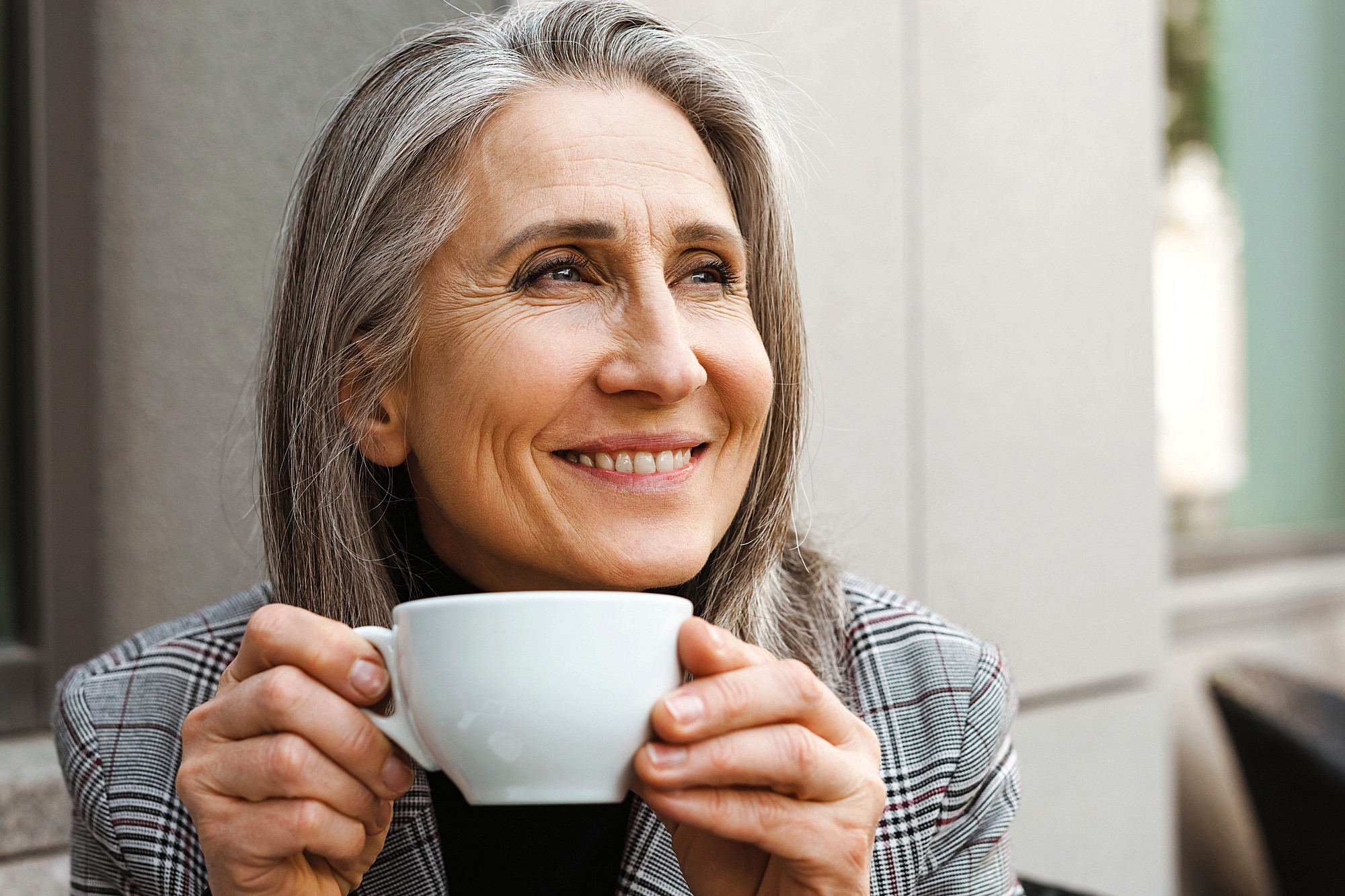 A smiling woman with long gray hair holds a white coffee cup, looking thoughtfully into the distance. She is wearing a plaid jacket and sitting outdoors, with a blurred building wall in the background.