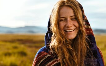 A smiling person with long red hair is wrapped in a colorful patterned blanket. They are standing outdoors in a grassy landscape under a cloudy sky, with mountains in the distance.