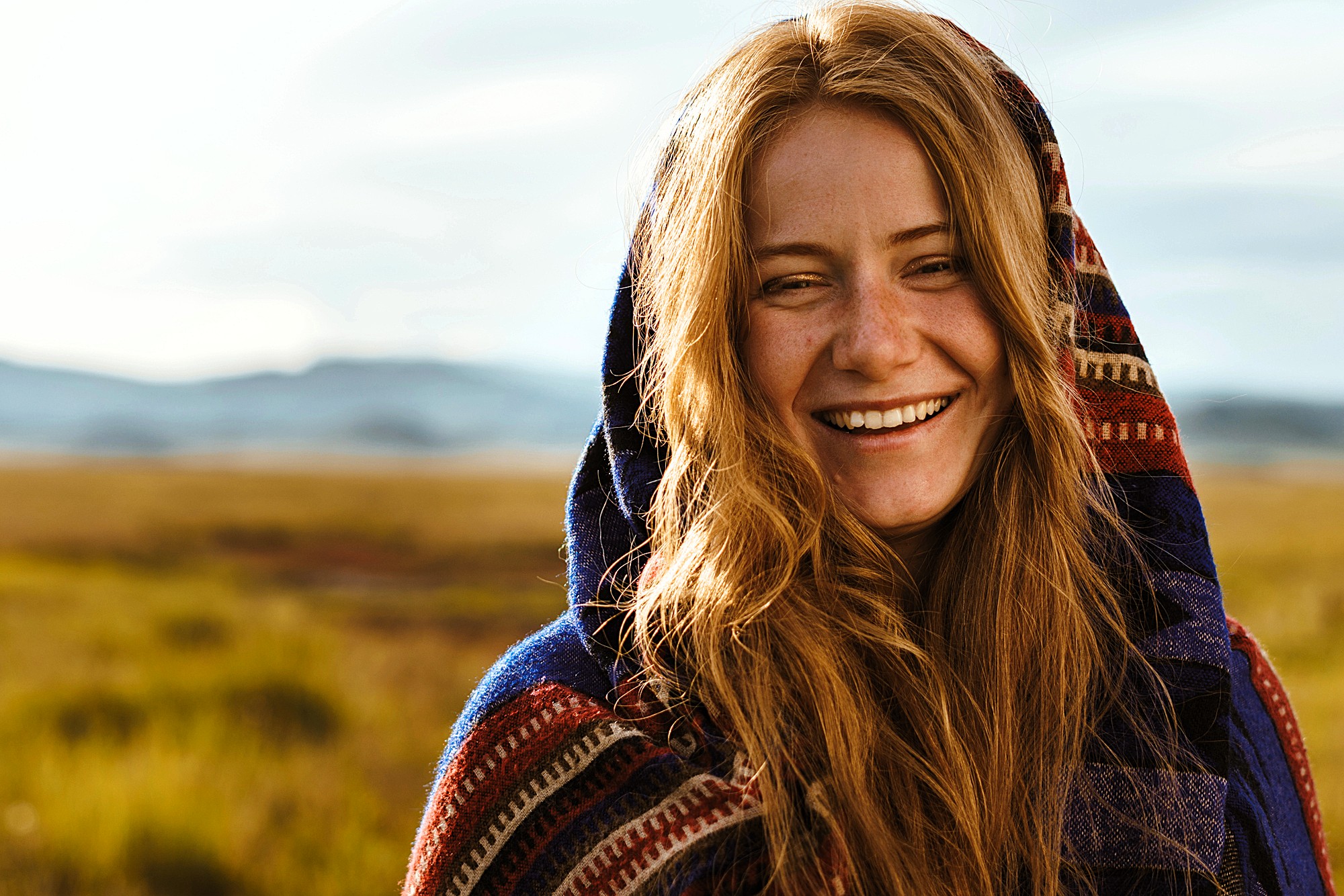 A smiling person with long red hair is wrapped in a colorful patterned blanket. They are standing outdoors in a grassy landscape under a cloudy sky, with mountains in the distance.