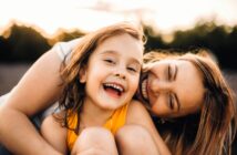 A young girl in a yellow top smiles and laughs while being hugged closely by a woman, likely her mother, outdoors. Both appear happy and joyful, with sunlight and trees in the blurred background.