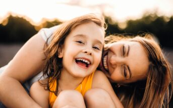A young girl in a yellow top smiles and laughs while being hugged closely by a woman, likely her mother, outdoors. Both appear happy and joyful, with sunlight and trees in the blurred background.