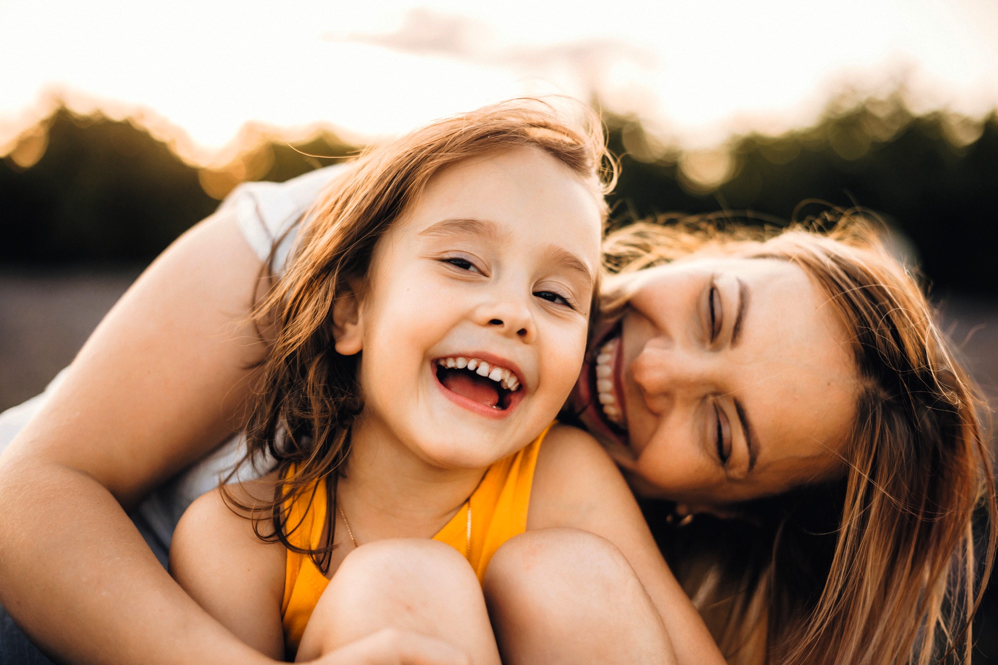 A young girl in a yellow top smiles and laughs while being hugged closely by a woman, likely her mother, outdoors. Both appear happy and joyful, with sunlight and trees in the blurred background.