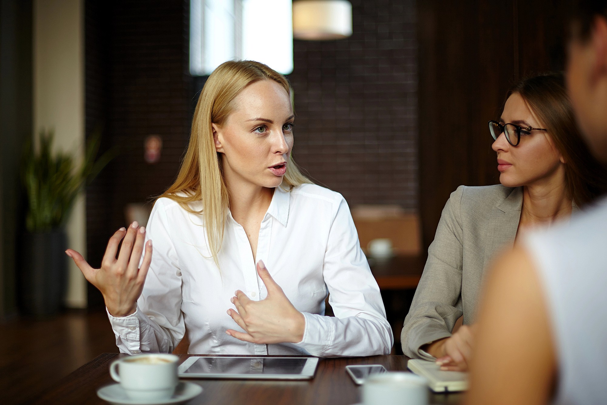 A woman in a white shirt is speaking and gesturing with her hands at a table, with a tablet in front of her. Another woman in glasses listens attentively. They are in a well-lit room with coffee cups on the table.