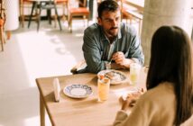 A man and a woman are sitting across from each other at a wooden table in a cafe, having a meal. The table has plates of food and two glasses of drinks. The man is looking attentively at the woman as they converse. The setting is bright and casual.