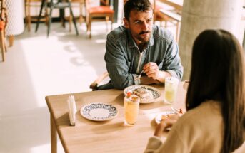 A man and a woman are sitting across from each other at a wooden table in a cafe, having a meal. The table has plates of food and two glasses of drinks. The man is looking attentively at the woman as they converse. The setting is bright and casual.