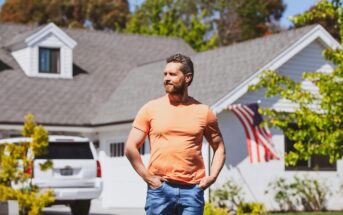 A man wearing an orange t-shirt and blue jeans stands outside with his hands in his pockets, in front of a white house with an American flag and a white SUV parked in the driveway. Green trees surround the sunny scene.