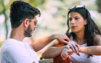 Two people sit outdoors, engaged in a conversation. The man, wearing sunglasses and a white shirt, looks at the woman, who is also wearing white and holding her arm, with sunglasses resting on her head. The background is blurred greenery.