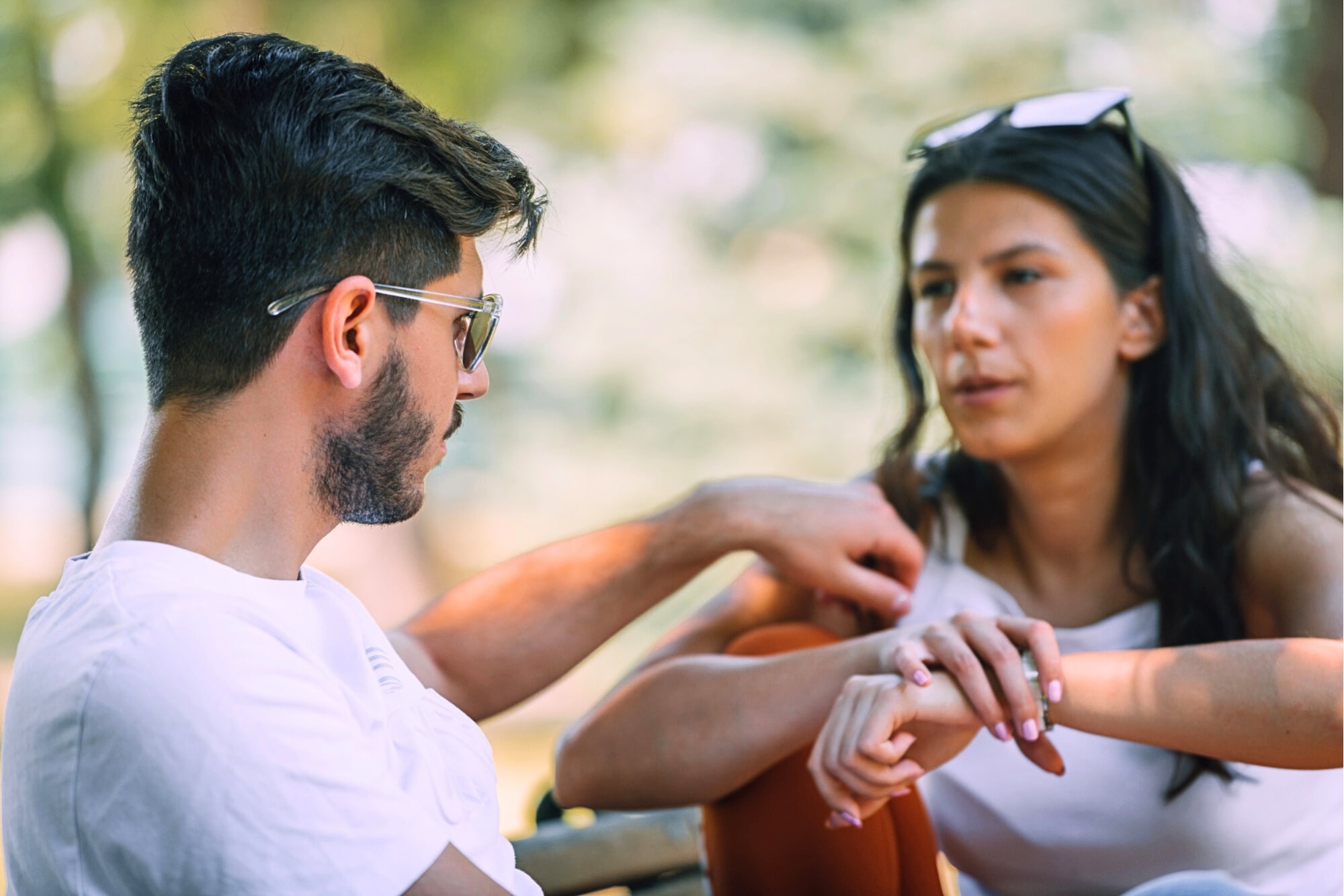 Two people sit outdoors, engaged in a conversation. The man, wearing sunglasses and a white shirt, looks at the woman, who is also wearing white and holding her arm, with sunglasses resting on her head. The background is blurred greenery.