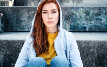 A young woman with long red hair, wearing a light gray hoodie and yellow shirt, sits on concrete steps outdoors, looking directly at the camera with a neutral expression.