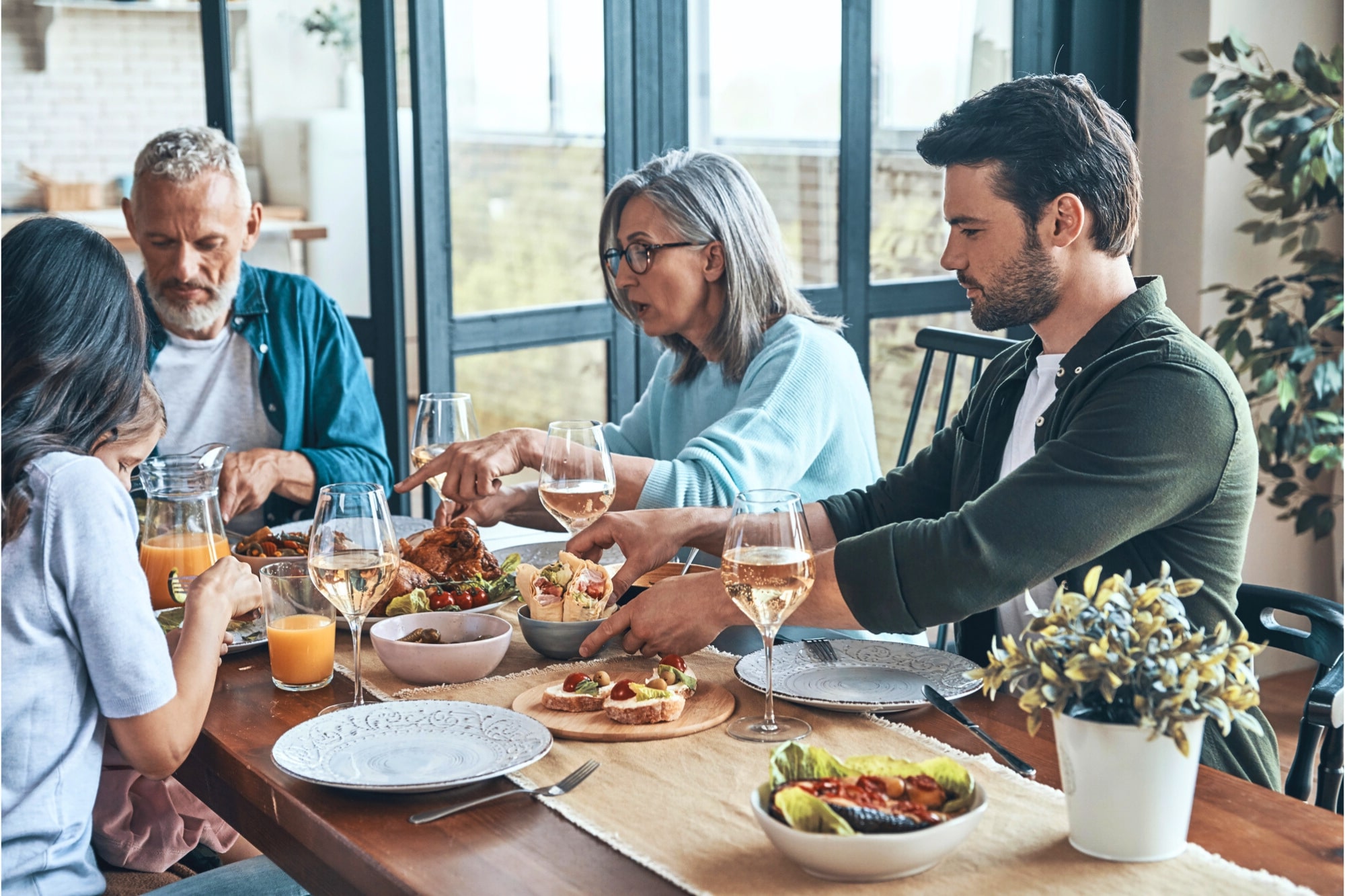 Four people sit around a dining table sharing a meal, with plates of food, glasses of wine and juice, and a potted plant visible. They are engaged in conversation in a bright, modern room.