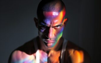 A man with a shaved head and intense expression sits in a white tank top. Colorful, rainbow light patterns are cast across his face and shoulders against a dark background.