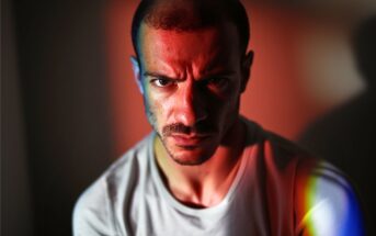 A man with a serious expression is lit by dramatic red and blue lighting, casting strong shadows across his face. He wears a light-colored shirt and looks directly at the camera.