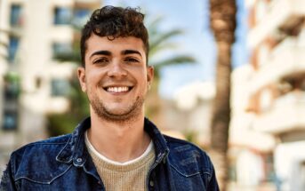 A young man with short curly hair and a light beard smiles outdoors, wearing a denim jacket over a beige sweater. The background shows blurred buildings, palm trees, and a bright blue sky.