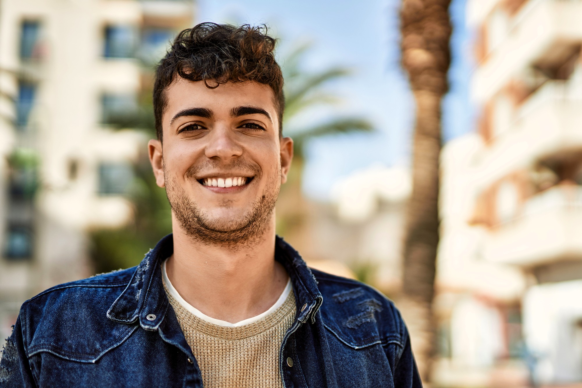 A young man with short curly hair and a light beard smiles outdoors, wearing a denim jacket over a beige sweater. The background shows blurred buildings, palm trees, and a bright blue sky.