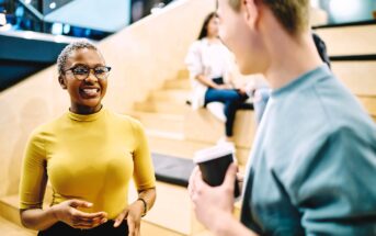 A woman in a yellow top and glasses smiles while talking to a man holding a coffee cup. They are standing in a modern, indoor setting with wooden steps and people in the background.
