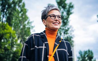A smiling older woman with short gray hair and glasses stands outdoors, wearing an orange turtleneck and a dark plaid coat. Green trees and a cloudy sky are in the background.