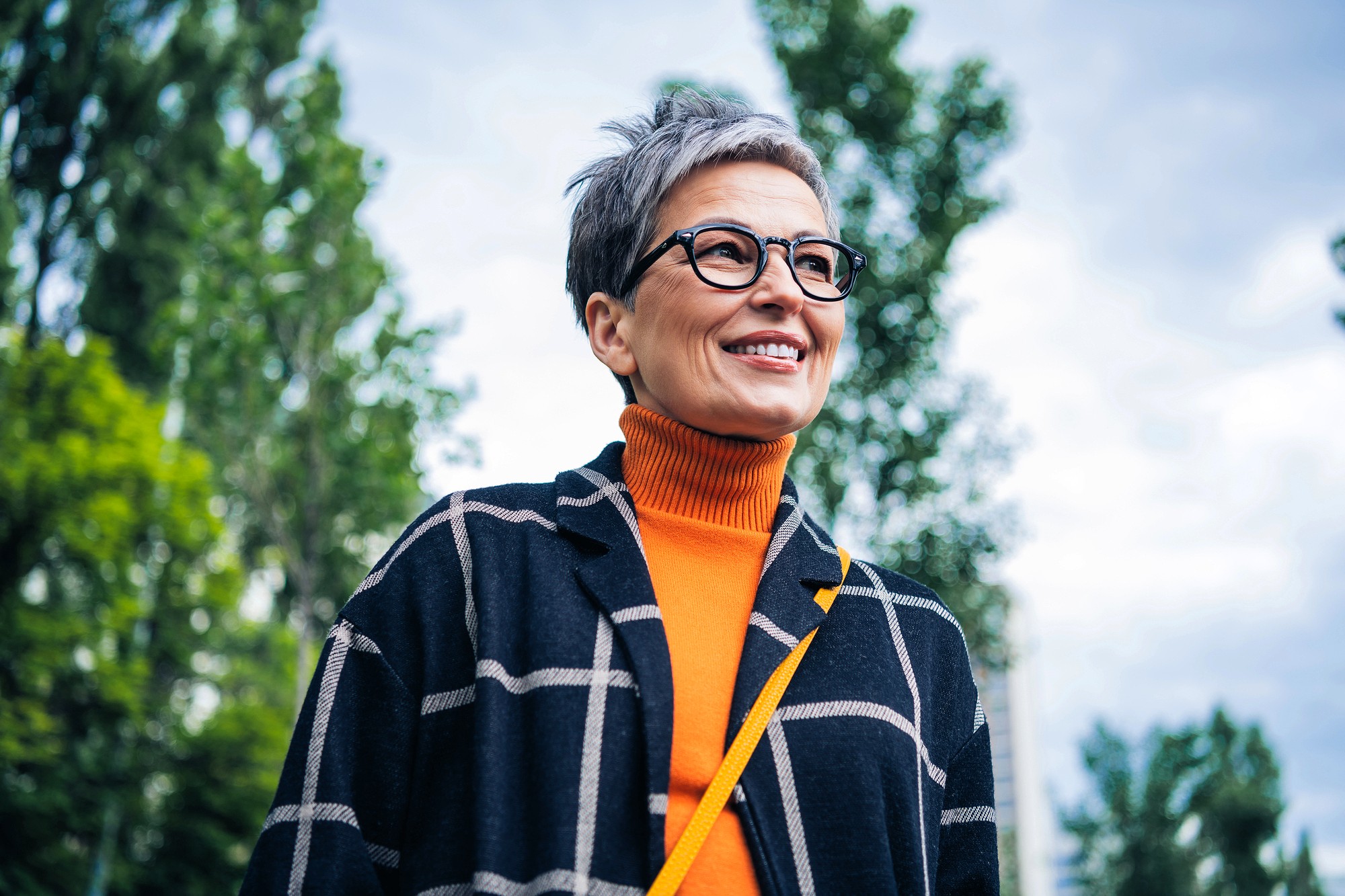 A smiling older woman with short gray hair and glasses stands outdoors, wearing an orange turtleneck and a dark plaid coat. Green trees and a cloudy sky are in the background.