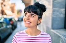 A young woman with dark hair in a bun, wearing a white and pink striped shirt, smiles while standing outside on a sunny day near a stone wall and blurred city street.