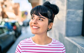A young woman with dark hair in a bun, wearing a white and pink striped shirt, smiles while standing outside on a sunny day near a stone wall and blurred city street.