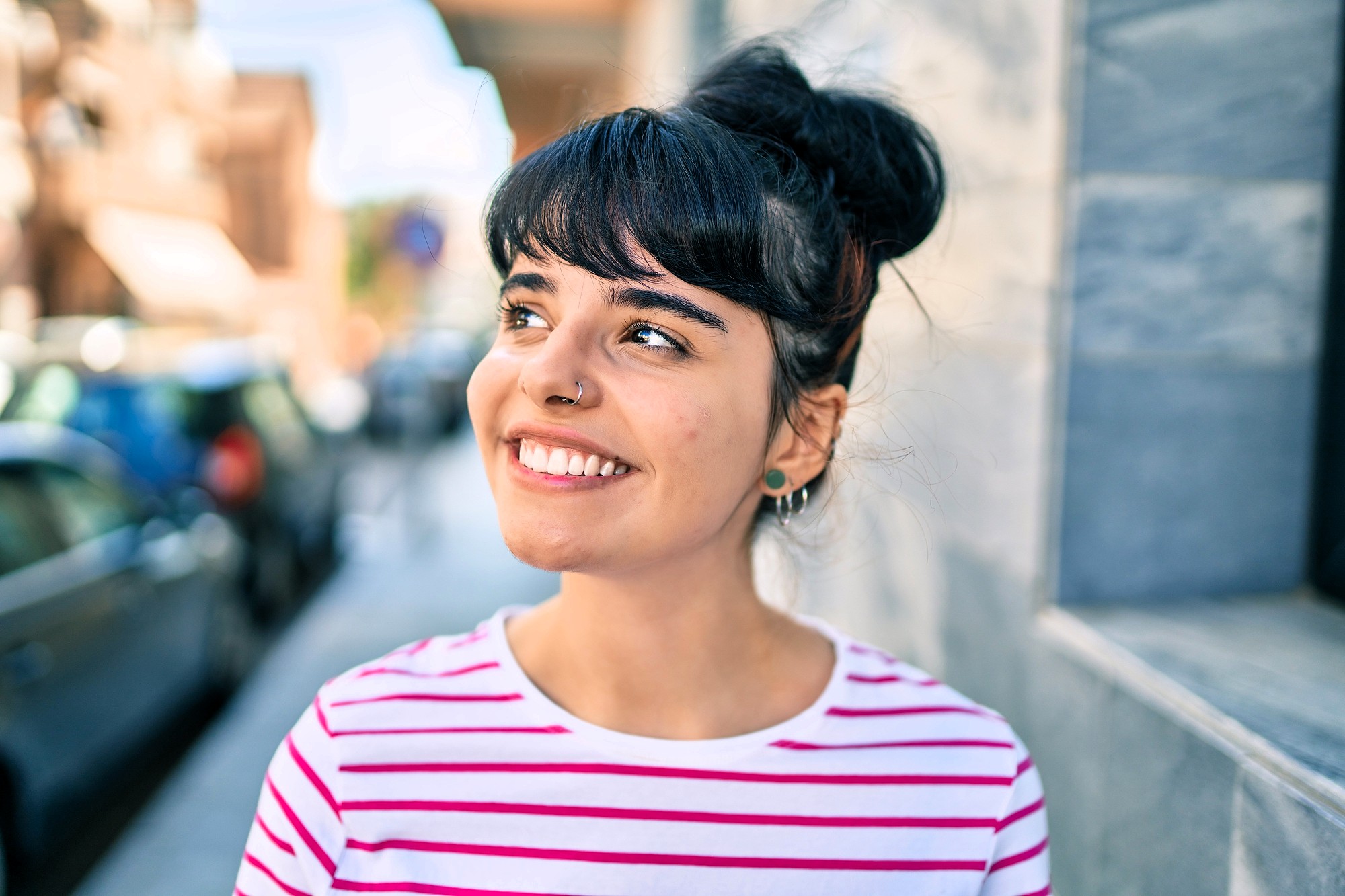A young woman with dark hair in a bun, wearing a white and pink striped shirt, smiles while standing outside on a sunny day near a stone wall and blurred city street.