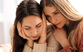 Two women sitting close together, one looking distressed with her head in her hands, while the other leans in, appearing to comfort and support her.