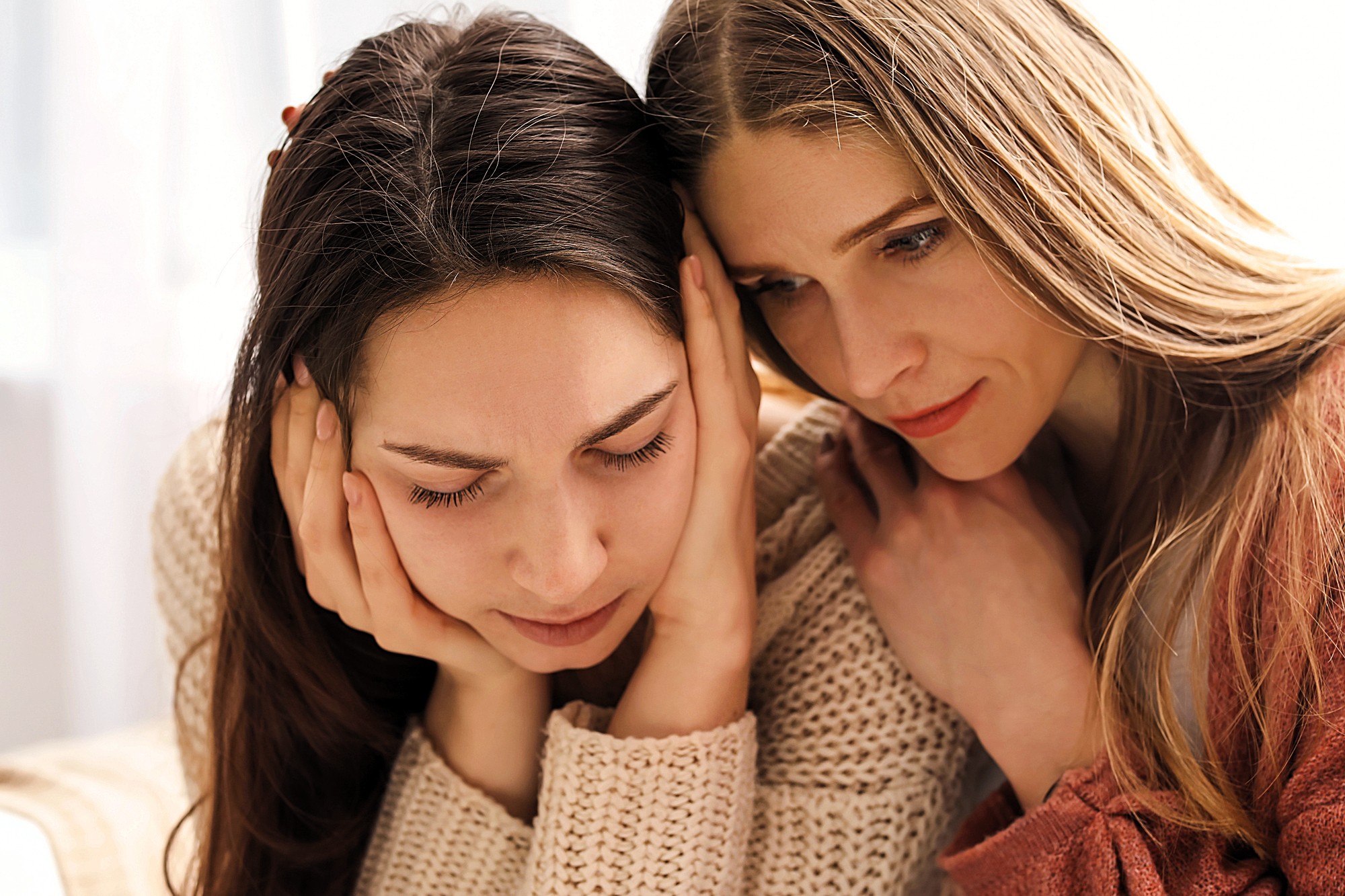 Two women sitting close together, one looking distressed with her head in her hands, while the other leans in, appearing to comfort and support her.