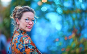 A woman with curly hair, wearing round glasses and a colorful, floral-patterned orange jacket, stands outdoors against a blurred blue and green leafy background.