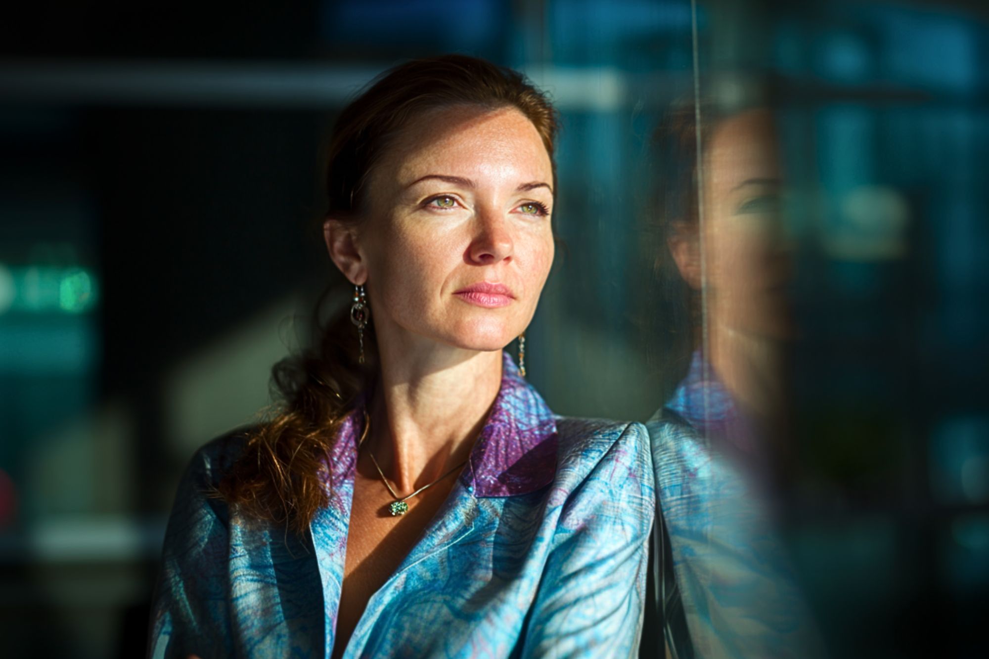 A woman in a blue patterned blazer gazes thoughtfully out a window, with sunlight illuminating her face and her reflection visible in the glass beside her.