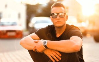A young man wearing sunglasses and a black t-shirt sits outdoors on the pavement with his arms resting on his knees. The sun is shining brightly behind him, creating a warm, glowing background.