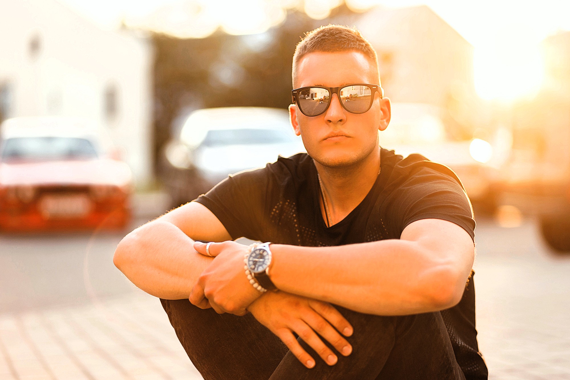 A young man wearing sunglasses and a black t-shirt sits outdoors on the pavement with his arms resting on his knees. The sun is shining brightly behind him, creating a warm, glowing background.