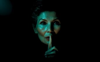 A woman with short hair holds a finger to her lips in a "shh" gesture, her face dramatically lit in blue-green light against a dark background, creating a mysterious atmosphere.